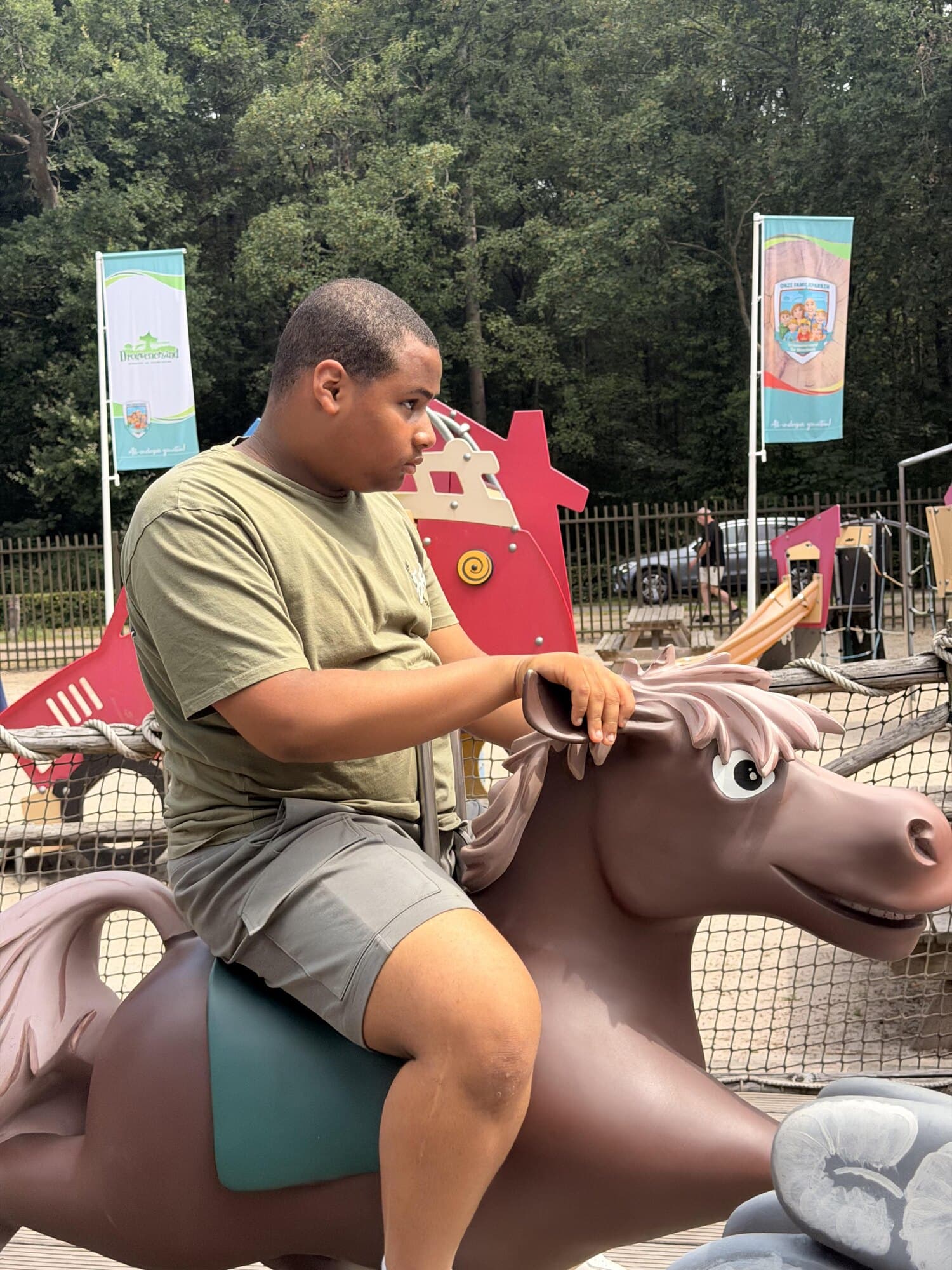 Een jongere zit op een kunststof speelpaard op het terrein van een attractiepark, met een hand op de manen. Achter het hek staan parkvlaggen tussen de bomen.
