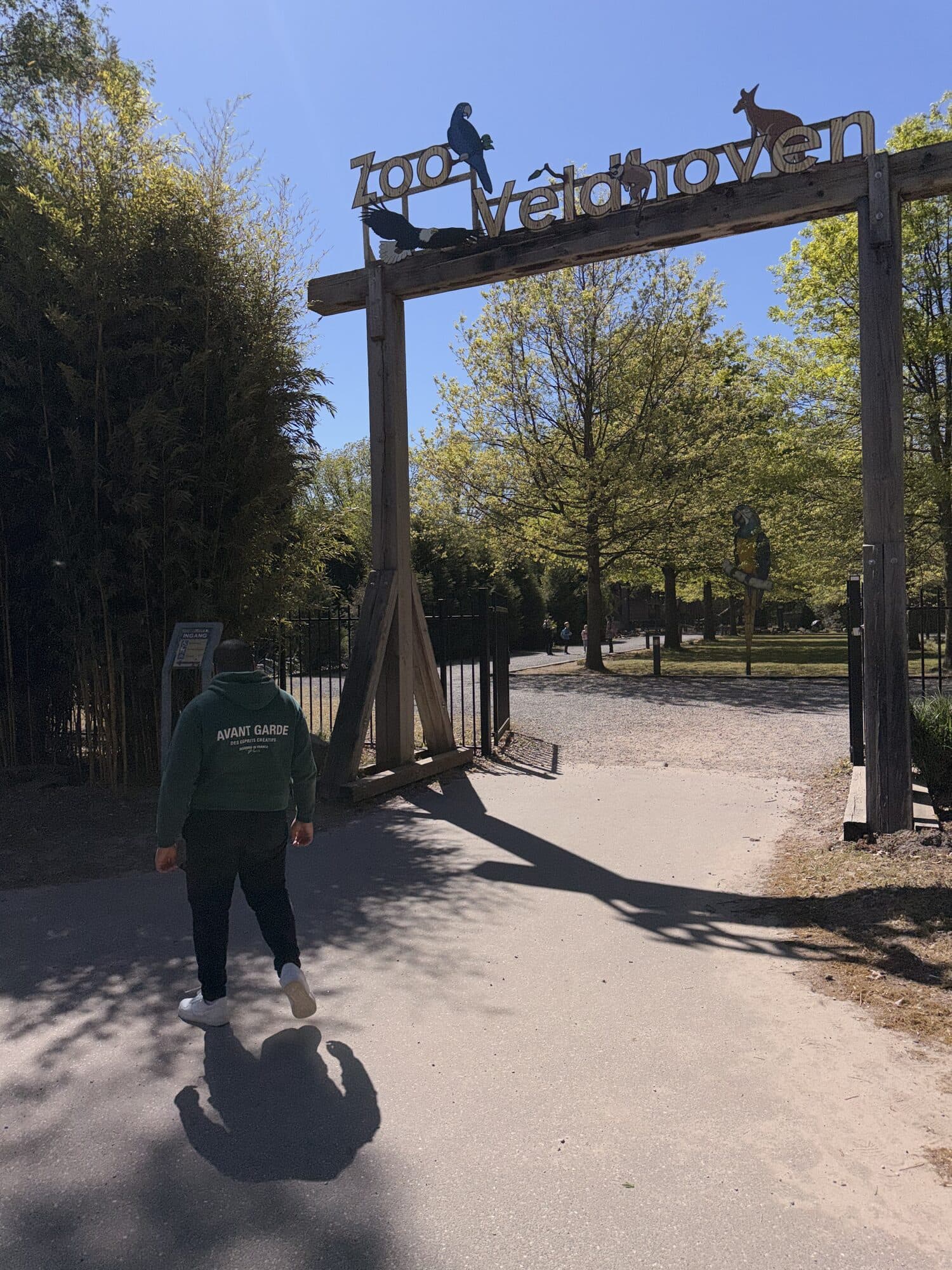 Een jongere loopt onder de houten toegangspoort van Zoo Veldhoven door, met bamboe langs het pad en de dierentuin achter het hek.