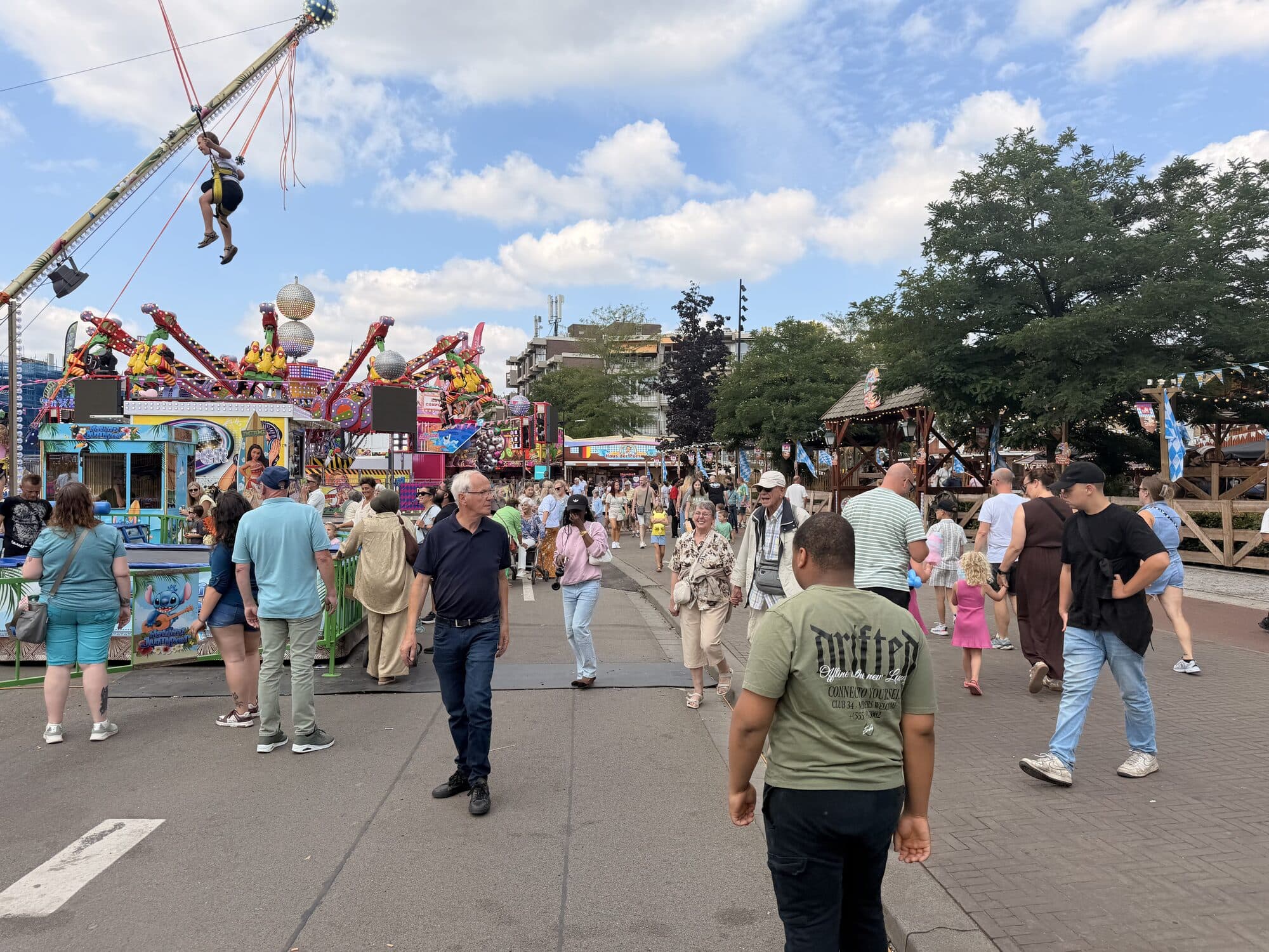 Drukke kermis met attracties, kramen en veel bezoekers onder een blauwe lucht met wolken.
