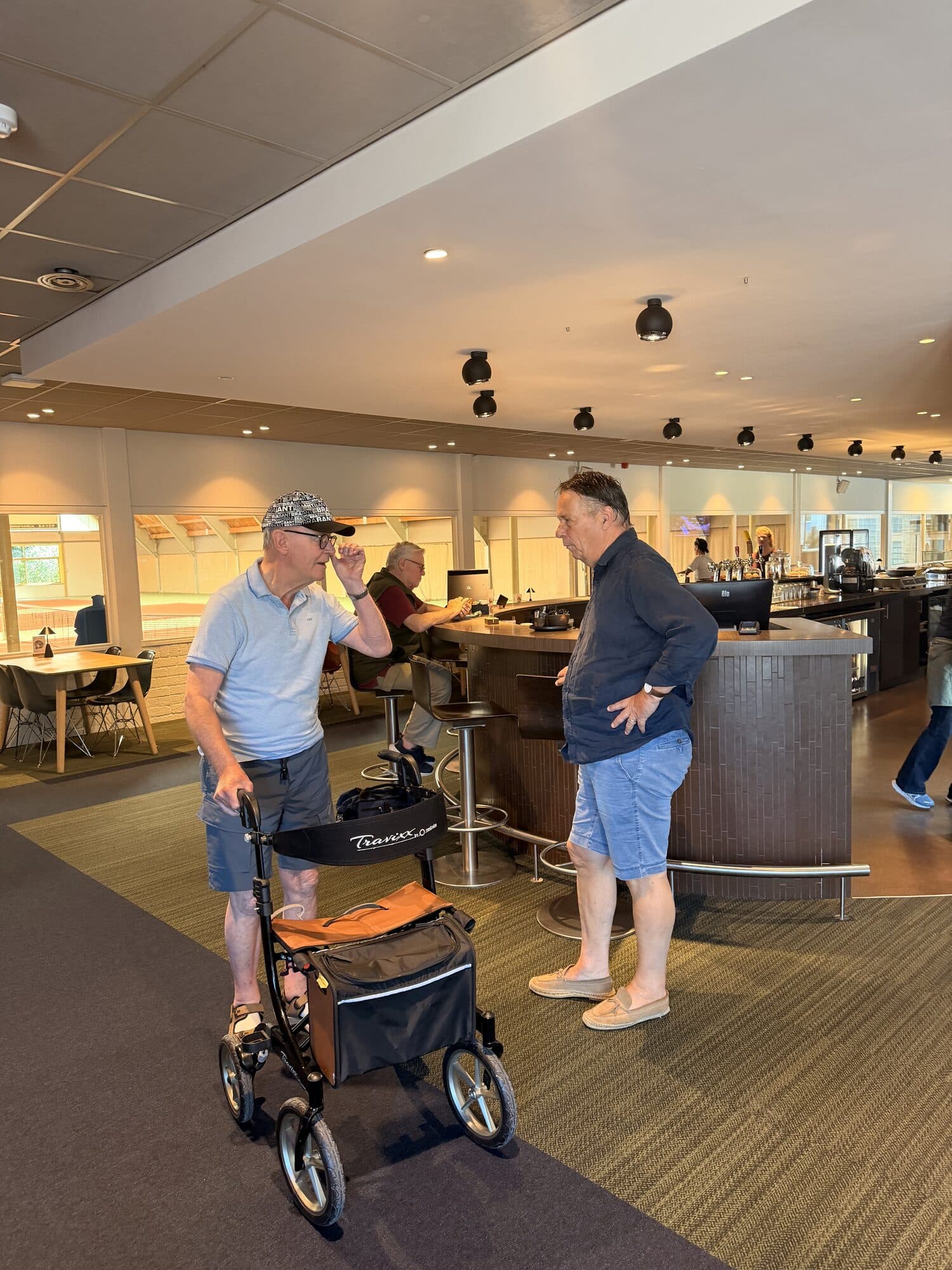 Twee mannen staan te praten in een winkelcentrum, een van hen met een rollator waar een tas aan hangt.
